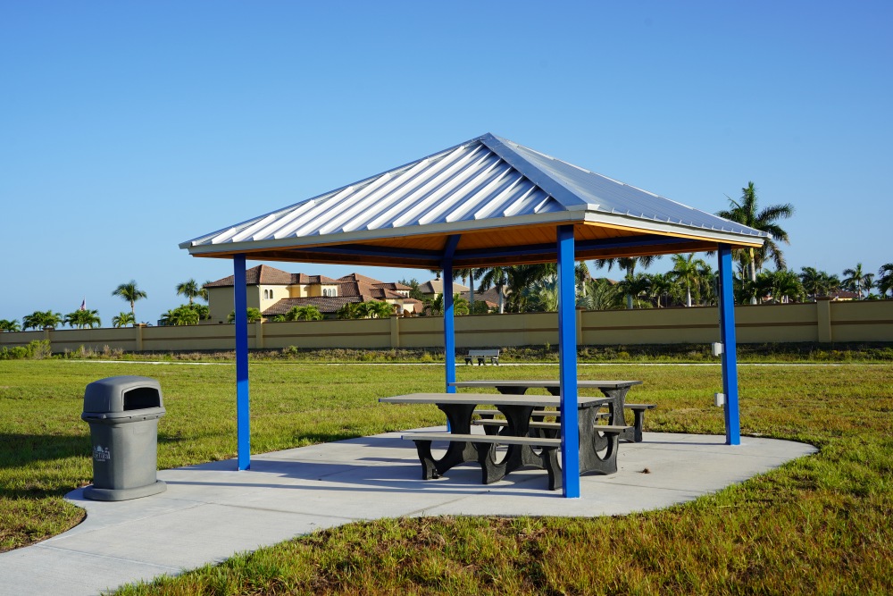 Picnic pavilion at Tropicana Park with a metal roof and blue posts, covering two picnic tables on a concrete pad. A trash can sits nearby on the lawn.